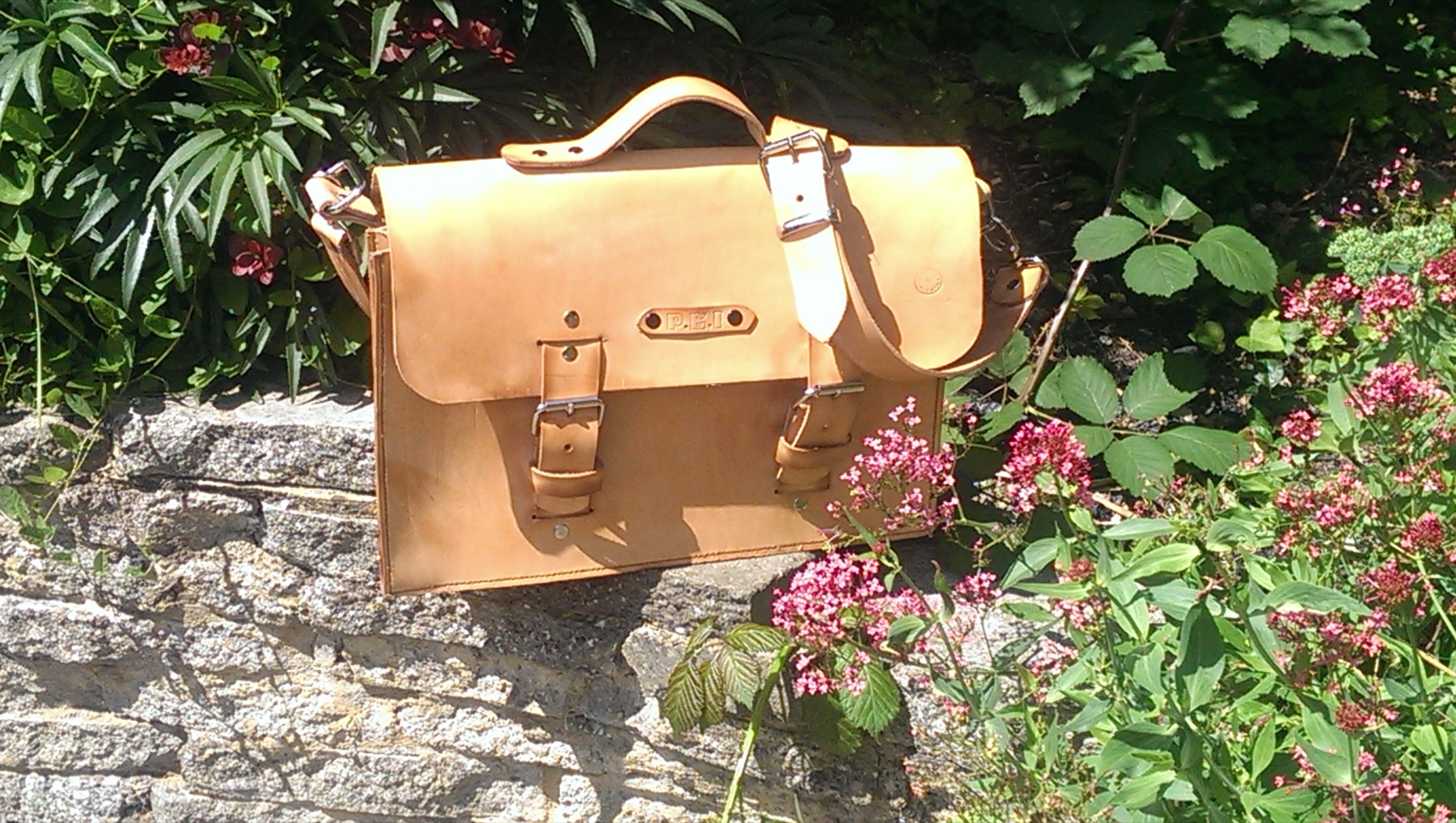A handmade tan leather satchel displayed outdoors among garden flowers